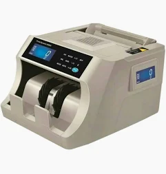 Mesin penghitung uang otomatis berwarna abu-abu muda dengan panel kontrol hitam di bagian depan. Memiliki layar digital LCD biru yang menunjukkan angka nol dan layar tambahan di sisi samping. Alat ini tampak kosong tanpa uang di dalamnya.